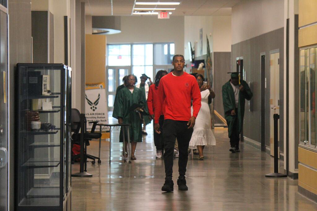 A person in a red shirt walks toward the camera in a school hallway while graduates in green gowns and an attendee in a white dress take photos in the background.