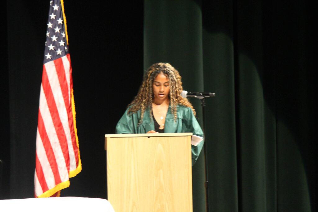 A person in a green graduation gown stands behind a podium onstage, reading from a paper with a microphone nearby. A U.S. flag stands beside the podium against dark green curtains.