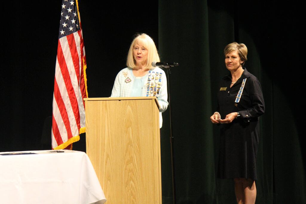 A person in a light blue dress stands at a podium speaking into a microphone, with a U.S. flag beside them and a musician with a double bass visible in the background.