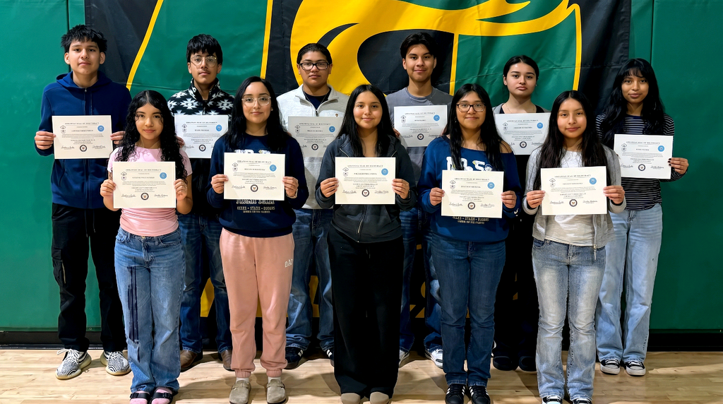 Group photo of eleven Mills University Studies High School students standing in two rows on a gym floor, each holding a Seal of Biliteracy certificate. A large green‑and‑yellow Mills banner hangs behind them. The front row has six students and the back row has five students, all facing the camera and displaying their certificates.