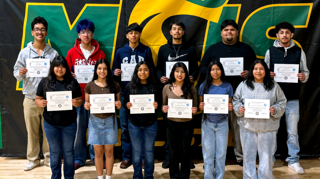 Group photo of twelve Mills University Studies High School students standing in two rows on a gym floor, each holding a Seal of Biliteracy certificate. A large green‑and‑yellow Mills banner hangs behind them. The front row has six students and the back row has five students, all facing the camera and displaying their certificates.