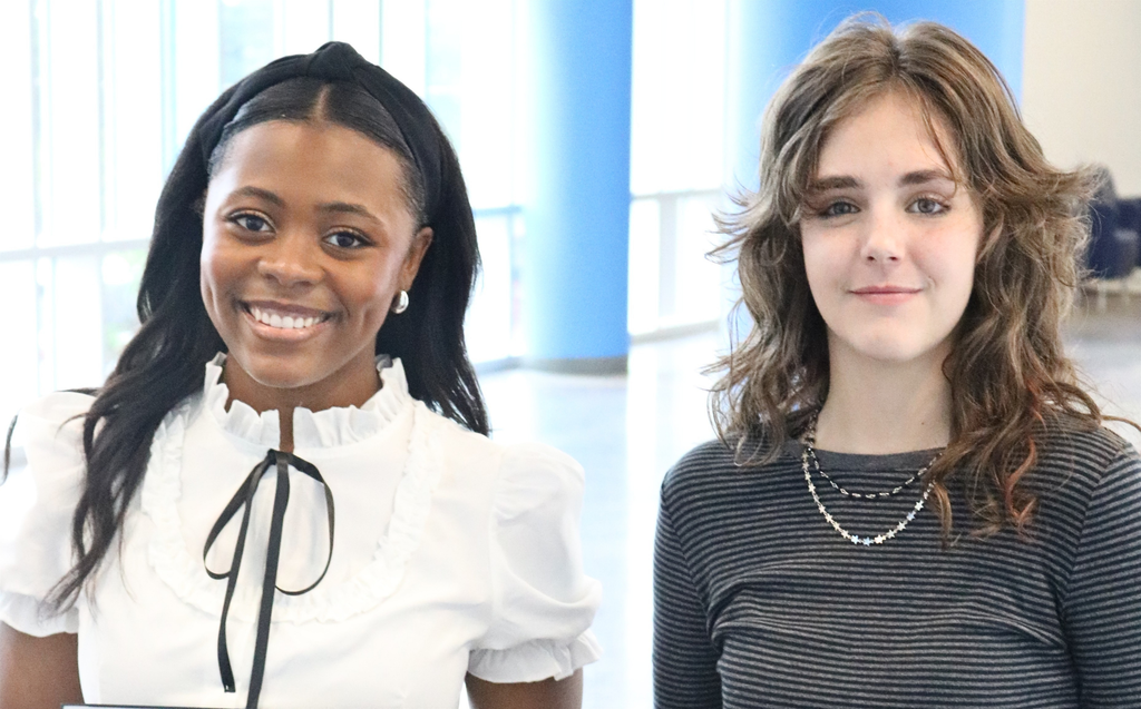 Morgan Perry and Hailey Middaugh stand side by side in a bright indoor space with large windows and blue columns behind them. Morgan, on the left, has long dark hair, is smiling, and wears a white blouse with a ruffled collar and black ribbon tie. Hailey, on the right, has wavy light brown hair, a neutral expression, and wears a dark striped shirt with layered necklaces.