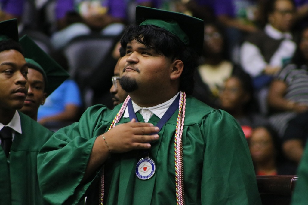 A graduate in a green cap and gown stands with a hand over their chest, wearing honor cords and a medal. Other graduates and a large audience are visible in the background.