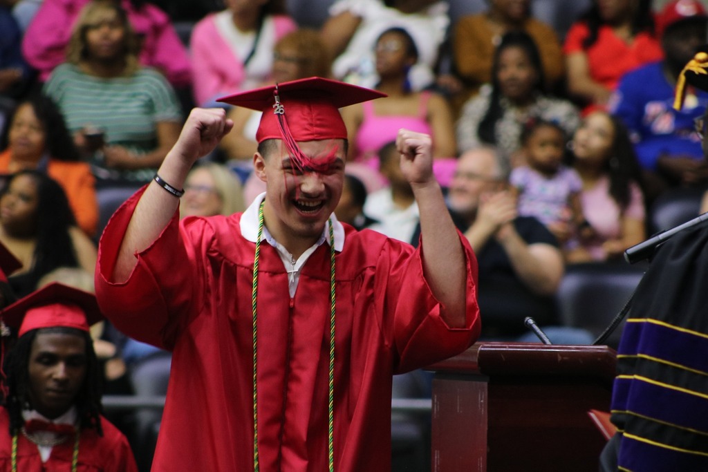A graduate in a red cap and gown raises both fists in celebration near a podium. Other red‑gown graduates and an audience are visible in the background.