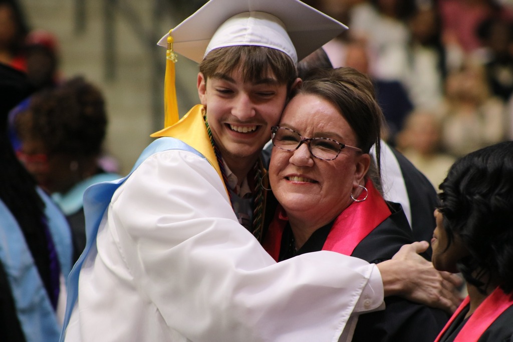 A graduate in a white cap and gown hugs a school board member wearing a black robe with a red collar. Other people stand nearby in a crowded graduation setting.