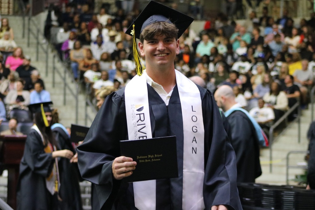  A graduate in a black cap and gown holds a diploma folder while standing in an indoor arena. The white stole reads “LOGAN” on one side and “DRIVEN SCHOOL OF INNOVATION” on the other. A large audience fills the bleachers in the background.