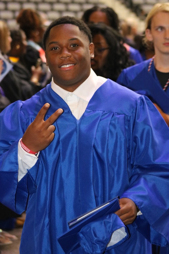 A graduate in a bright blue cap and gown holds a diploma folder in one hand and makes a peace sign with the other. Other blue‑gown graduates stand nearby in a large indoor venue.
