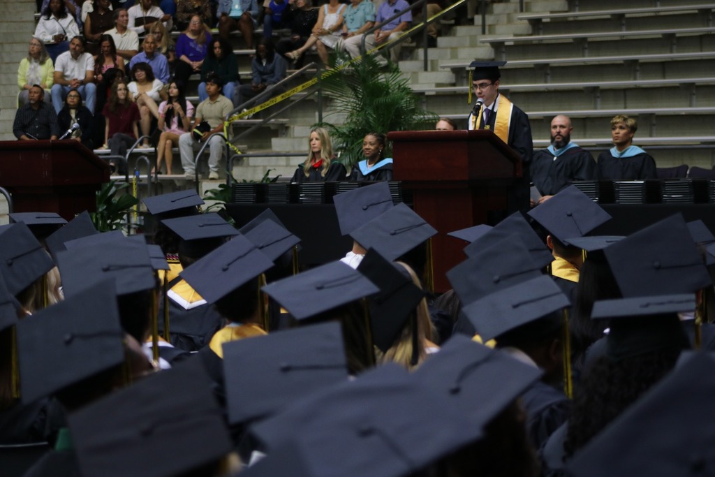 Alt text: A graduation ceremony in an indoor arena shows rows of graduates in black caps and gowns seated in front of a stage. A speaker in academic regalia stands at a podium while other officials sit behind them. Families fill the bleachers in the background.