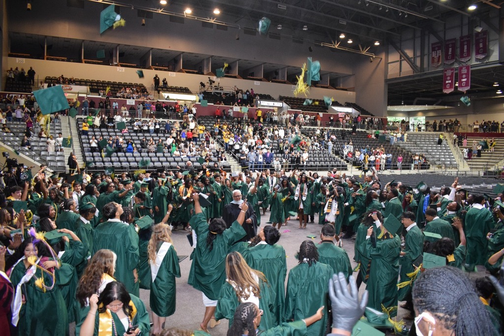 A large group of graduates in green caps and gowns toss their caps into the air inside an arena filled with cheering spectators.