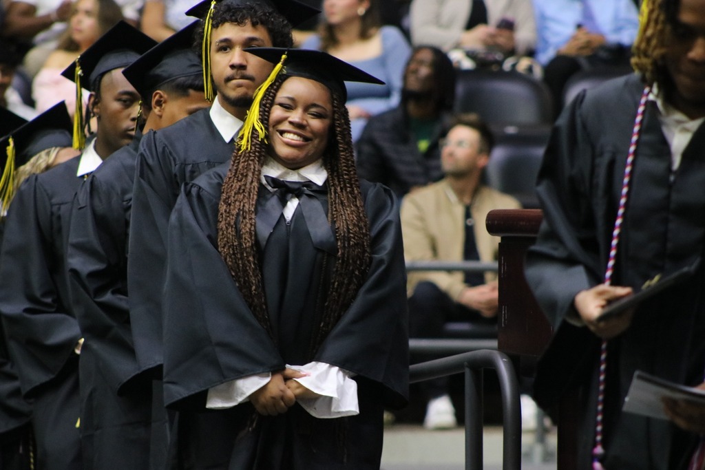 A group of graduates in black caps and gowns stand in line during a ceremony, with one graduate in the center smiling broadly as spectators watch.