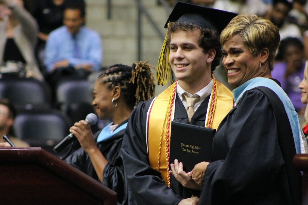 A graduate in a black cap and gown with honor cords holds a diploma folder labeled “Parkview High School, Little Rock, Arkansas” while standing beside an academic official on stage.