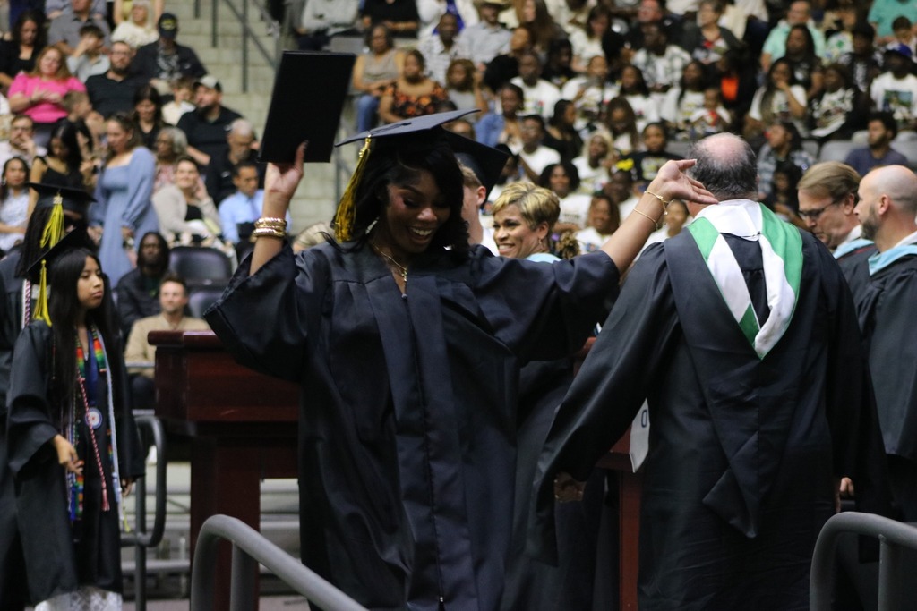 A graduate in a black cap and gown holds up a diploma folder while walking across the stage as faculty members congratulate students and the audience watches from the bleachers.