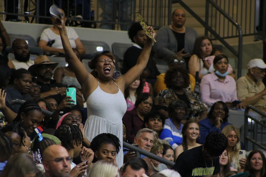 An audience member in a gray dress stands with both arms raised in celebration during a crowded indoor event as people around them clap and record on their phones.