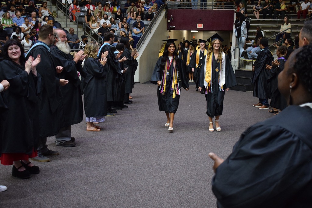 Two graduates in black caps and gowns walk down an aisle lined with clapping faculty members, with more graduates and spectators visible behind them.