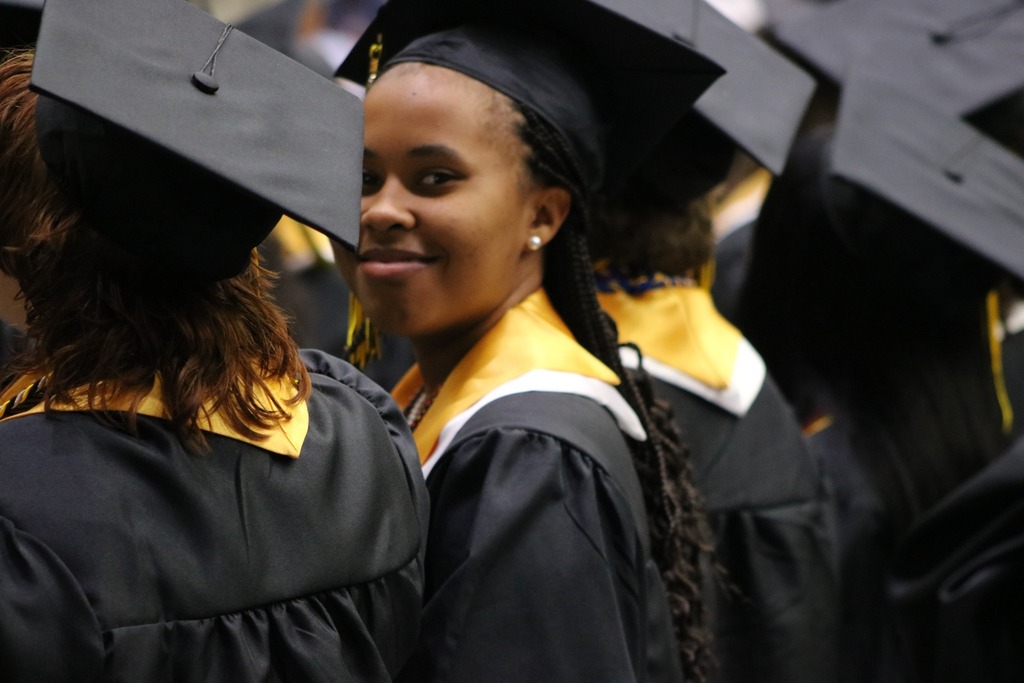 Graduates in black gowns and caps with gold stoles stand together during a ceremony, with one graduate turned slightly toward the camera.