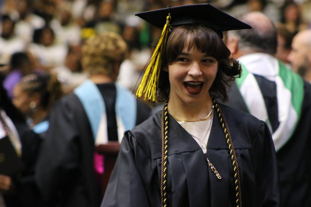 A graduate wearing a black cap with a yellow tassel and a black gown with honor cords stands among other graduates during an indoor ceremony.