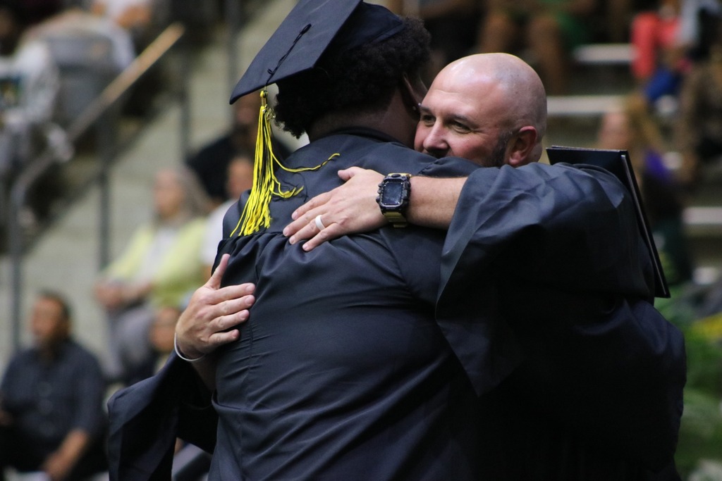 Two people in black caps and gowns hug during an indoor graduation ceremony, with seated guests visible in the background.