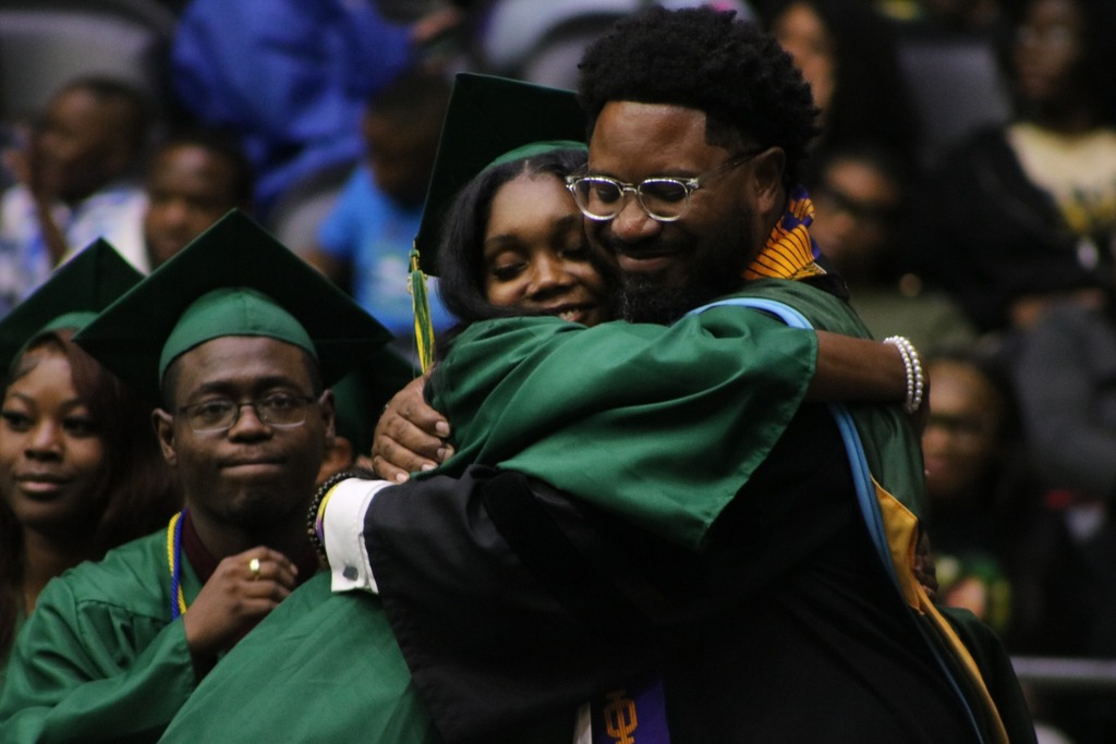 A graduate in a green cap and gown hugs a faculty member in colorful academic regalia while other graduates sit nearby.
