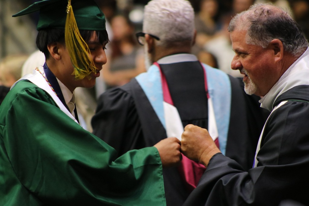 A graduate in a green cap and gown with a white stole gives a fist bump to an academic official during a graduation ceremony.