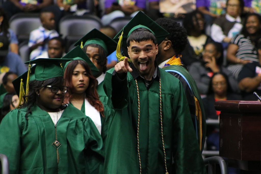  A graduate in a green cap and gown points forward and sticks out their tongue while standing with other graduates during a ceremony.