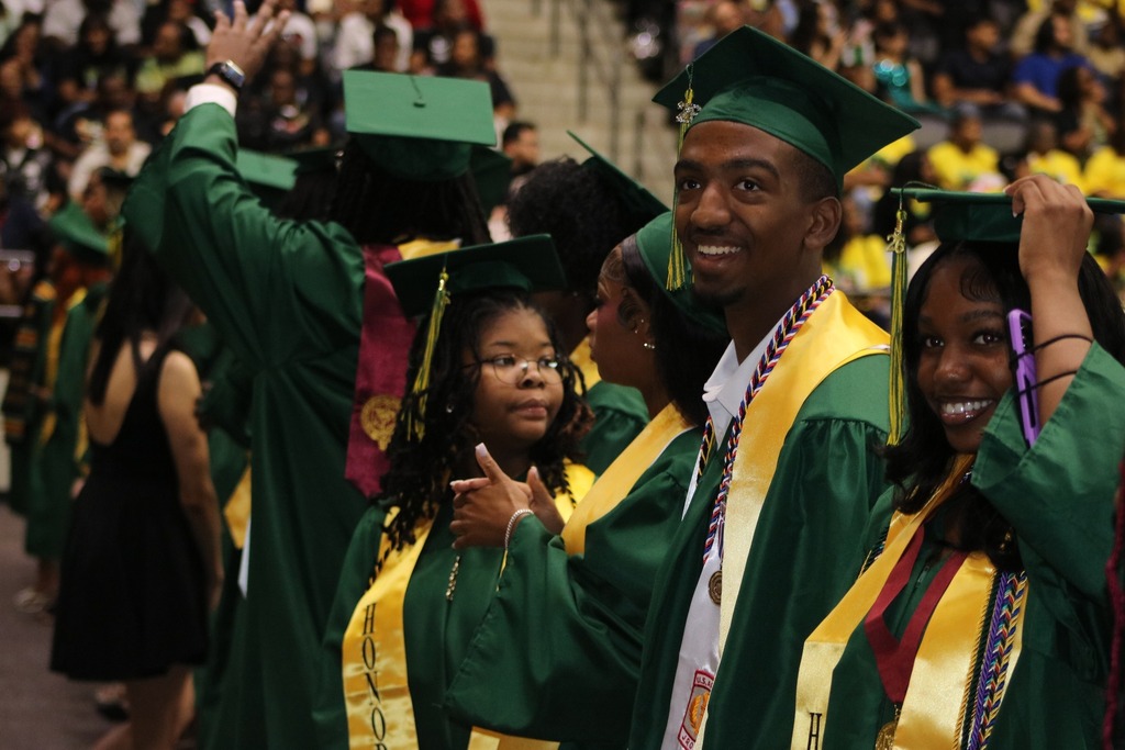 Graduates in green caps and gowns with yellow stoles stand in a line smiling as the audience watches from the bleachers.