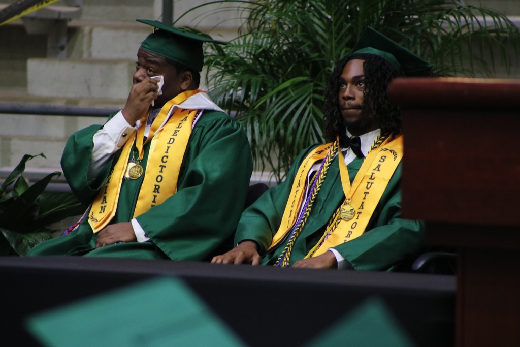 Two graduates in green caps and gowns sit on stage wearing stoles labeled “Valedictorian” and “Salutatorian,” with plants and a podium nearby.