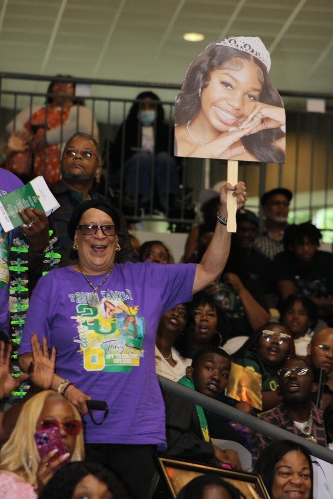 An audience member holds a large cutout photo of a graduate while seated among a cheering crowd at an indoor event.