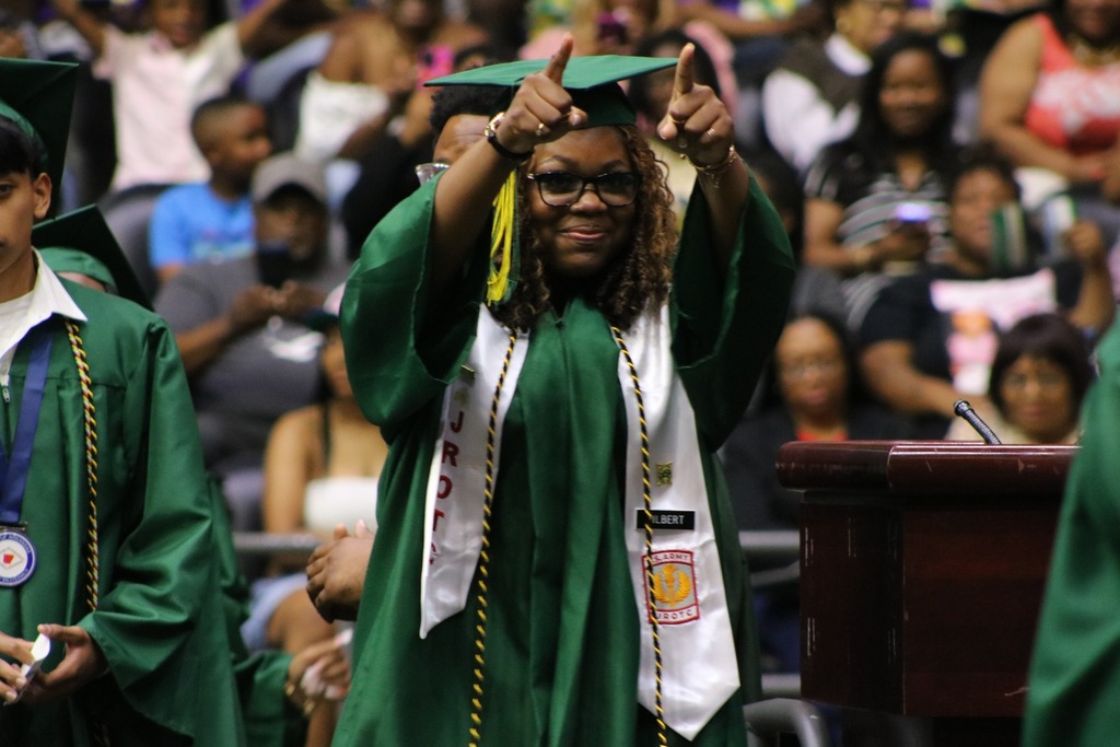 A graduate in a green cap and gown wearing a white JROTC stole raises both hands with index fingers pointing upward near a podium.