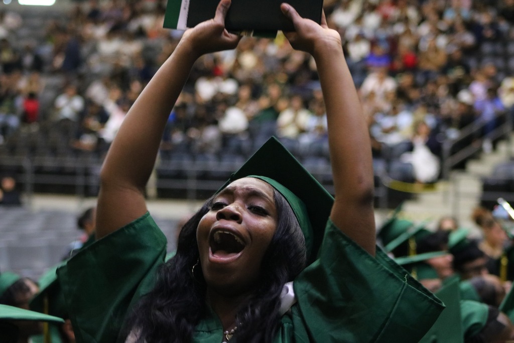 A graduate in a green cap and gown raises a diploma folder above their head in celebration as the audience watches.
