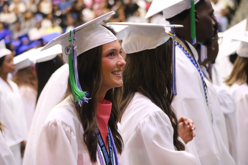 A graduate in a white cap and gown smiles while standing among classmates during a ceremony in a crowded indoor venue.