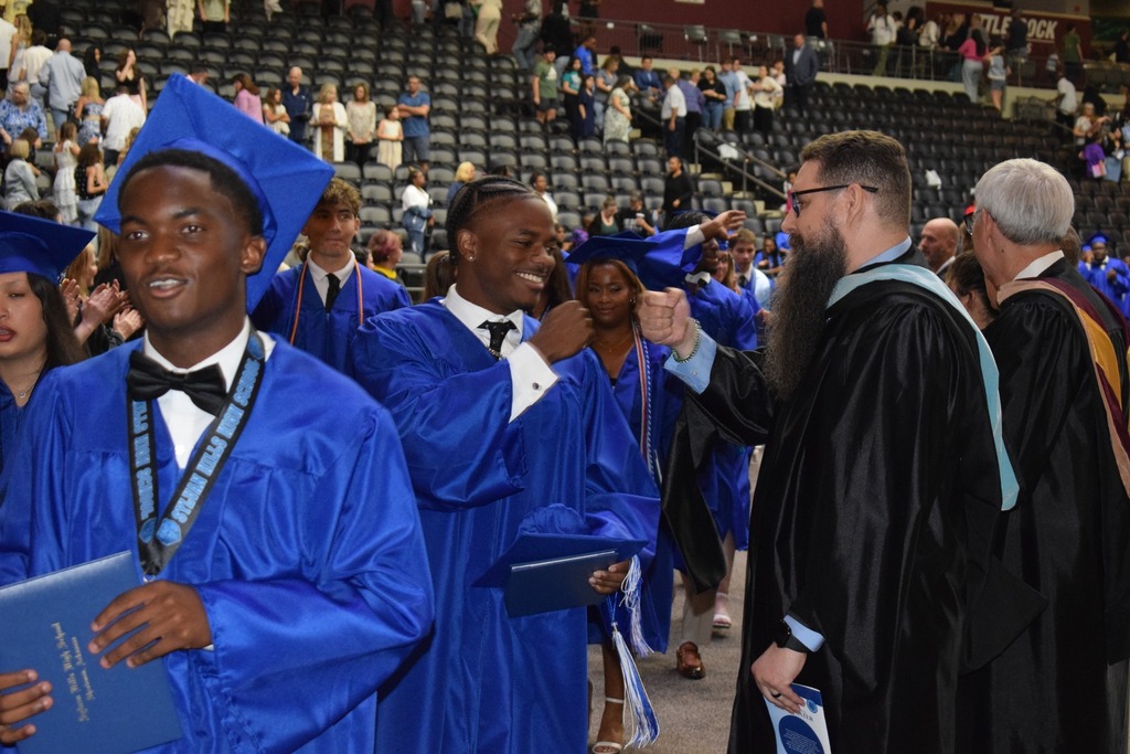 A graduate in a bright blue cap and gown gives a fist bump to a faculty member in academic regalia during a procession in an arena.