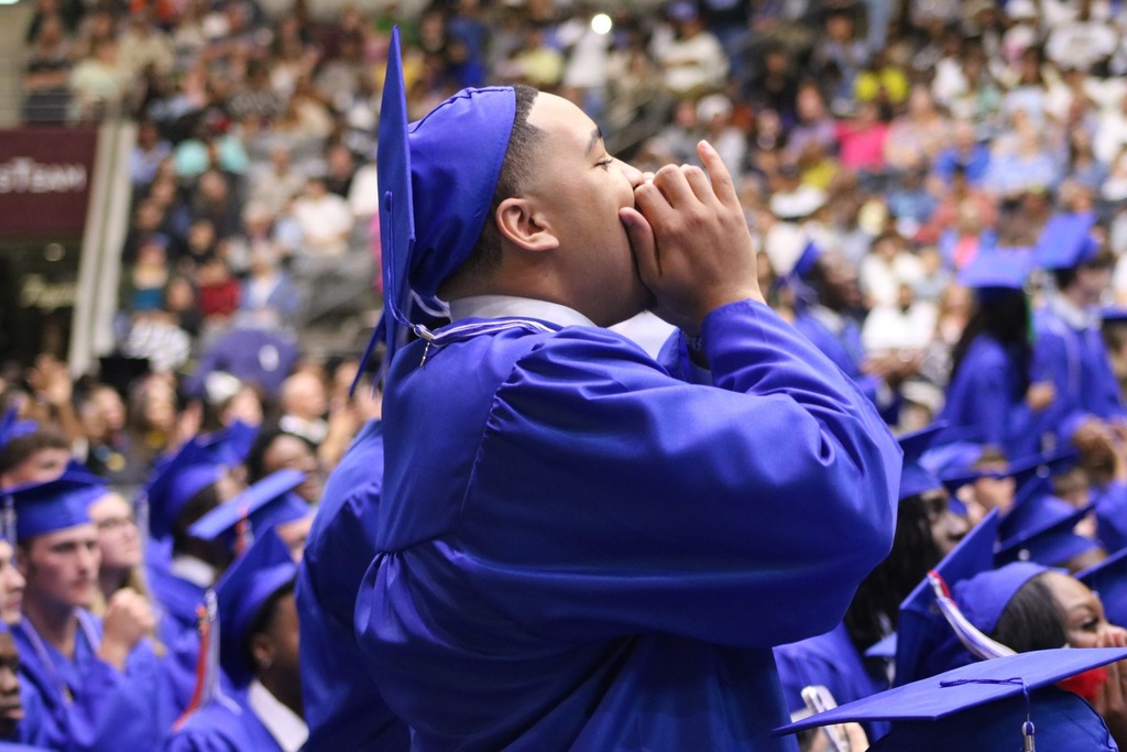 A graduate in a blue cap and gown stands and cheers with hands cupped around their mouth while other graduates sit nearby in an arena.