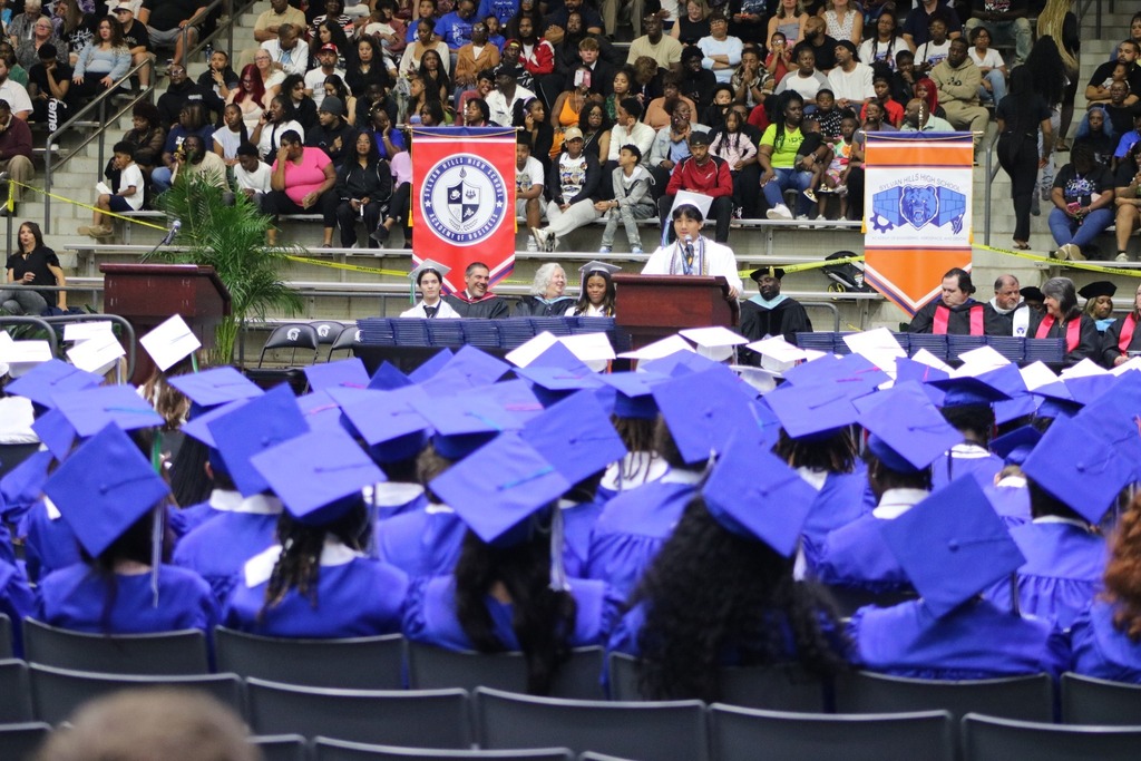 Graduates in blue caps and gowns sit facing a stage where a speaker stands at a podium, with banners and an audience visible in the background.