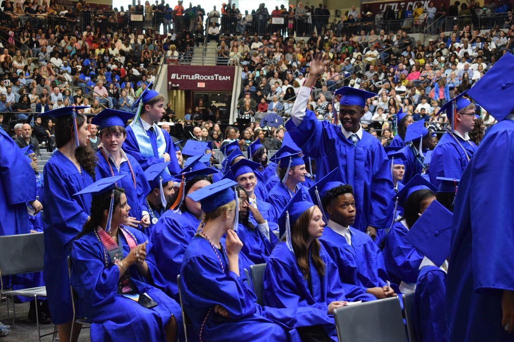 A graduate in a blue cap and gown stands and raises a hand while other graduates sit nearby and the audience fills the arena stands.