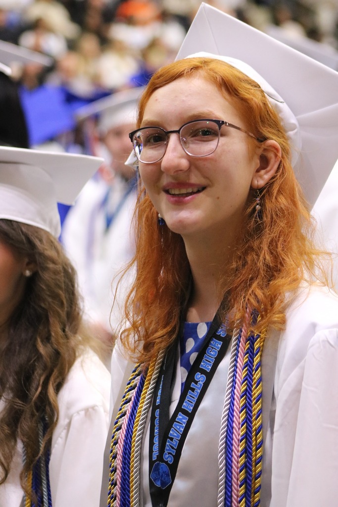 A graduate in a white cap and gown stands among classmates, wearing multiple honor cords and a lanyard, with other graduates visible in the background.