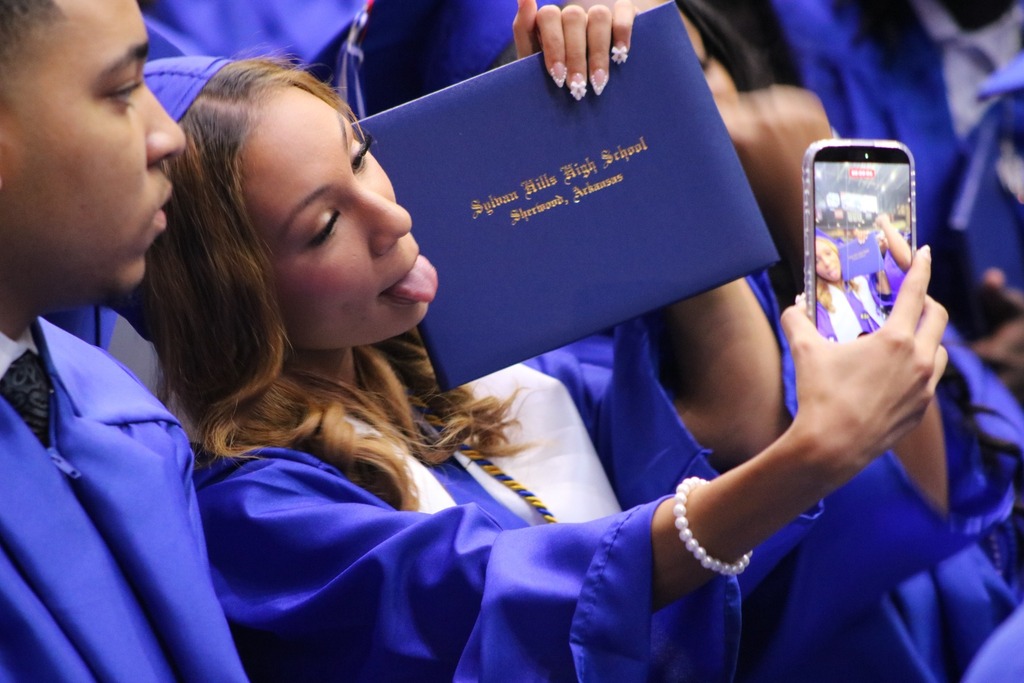  A graduate in a blue cap and gown takes a selfie while holding a diploma cover, with another graduate seated beside them.