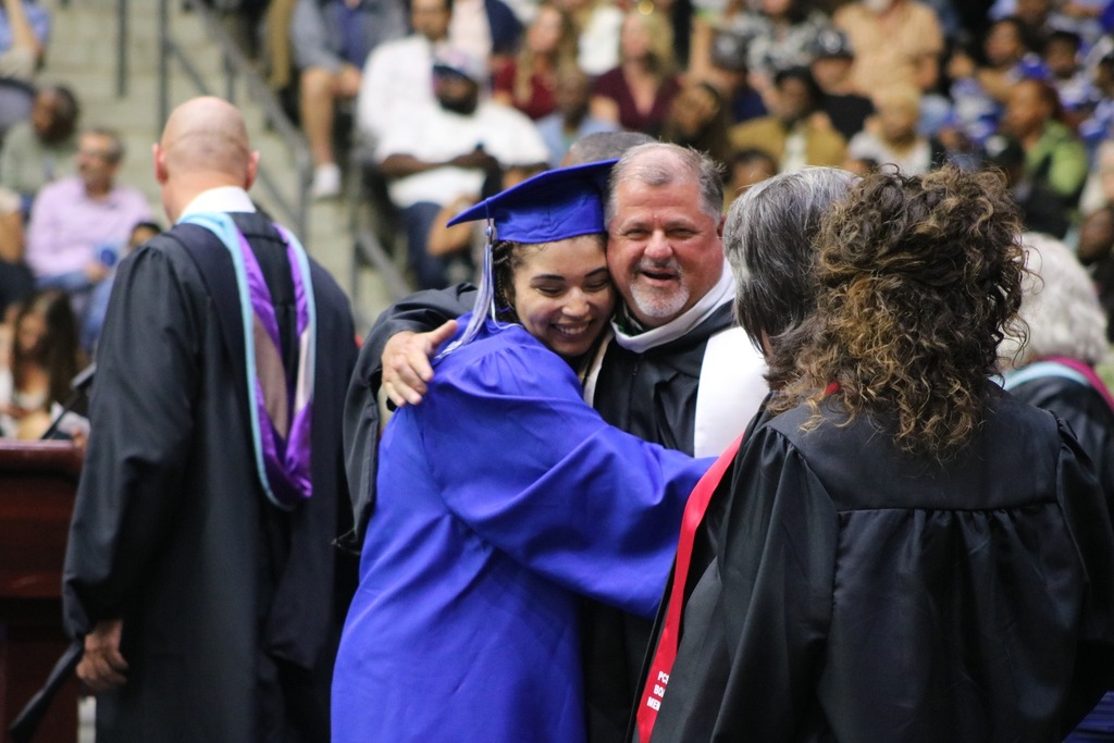 A graduate in a blue cap and gown hugs an academic official in a black robe with a white stole as other officials and the audience look on.