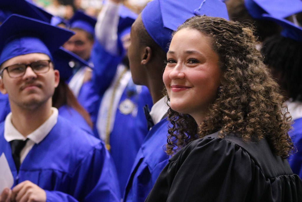 Graduates in blue caps and gowns gather closely during a ceremony, with one person in a black gown standing in the foreground.
