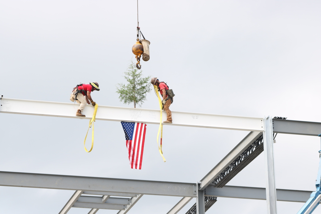 Construction workers stand atop a steel beam with an American flag and small tree attached before it is raised into place at Joe T. Robinson High School.