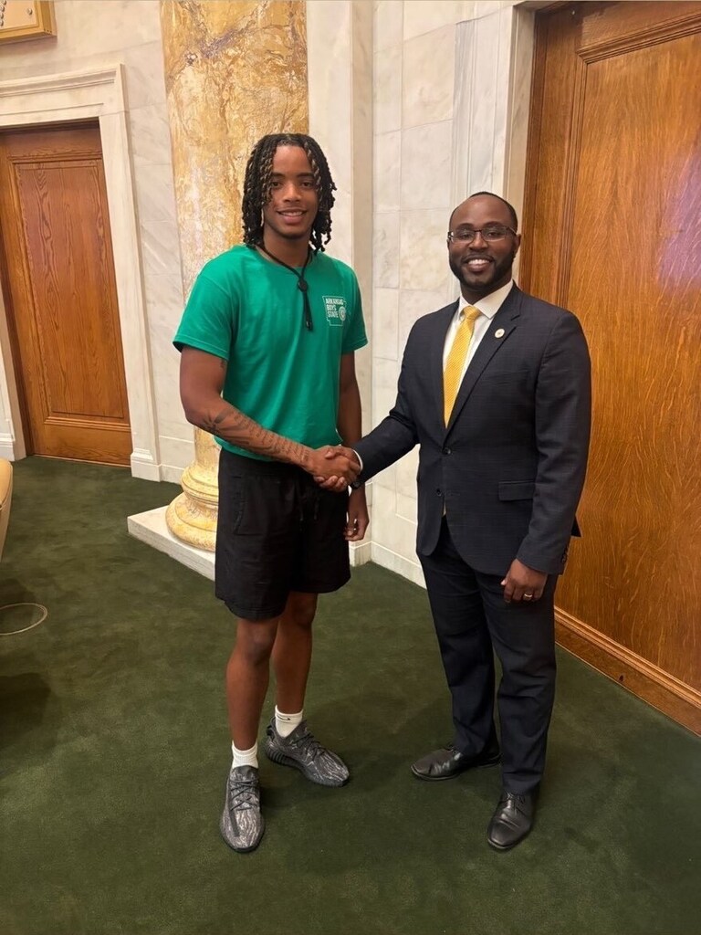Two individuals stand in a formal building with marble columns. One wears a green “Arkansas State University” T‑shirt; the other wears a dark suit and yellow tie. They shake hands and smile.