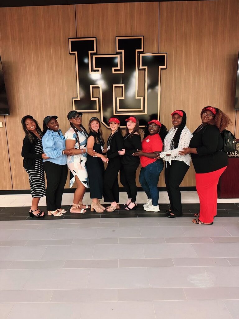 Eight people stand indoors in front of a large black‑and‑gold “HU” wall display. Several wear red visors and name badges, indicating participation in a leadership program.