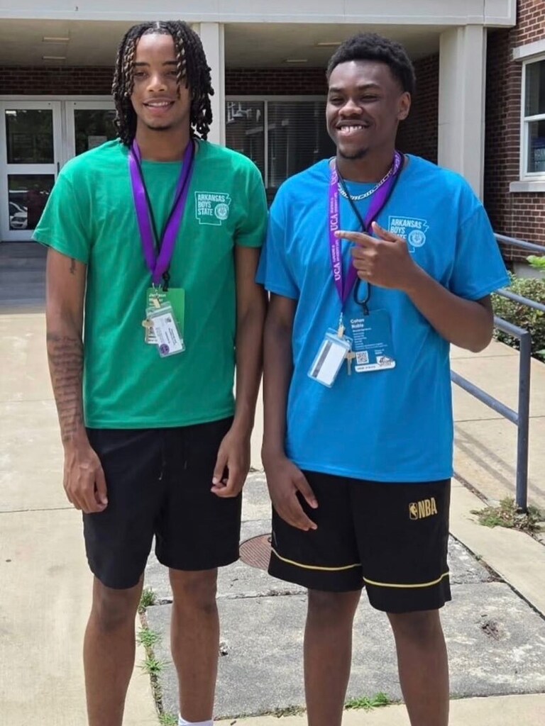 Two students stand outside a building with glass doors. One wears a green “Arkansas Boys State” T‑shirt; the other wears a blue version. Both have purple lanyards with ID badges.