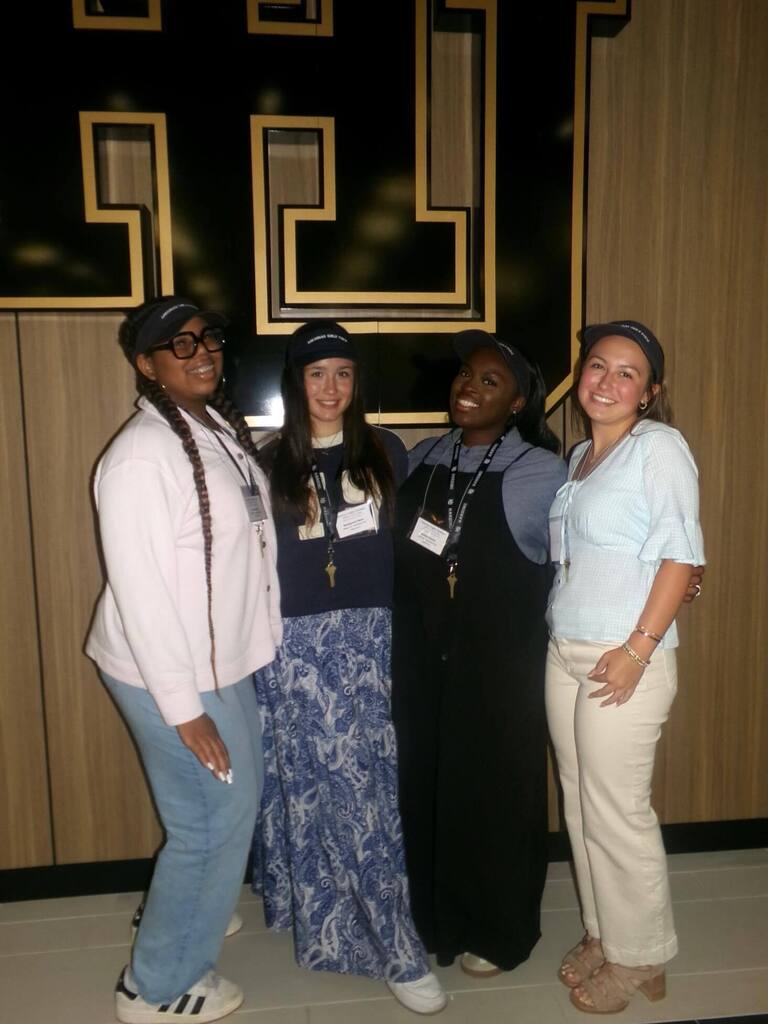 Four individuals stand indoors in front of a large black‑and‑gold “H” wall display. All wear dark caps, name badges, and lanyards, dressed in casual attire.