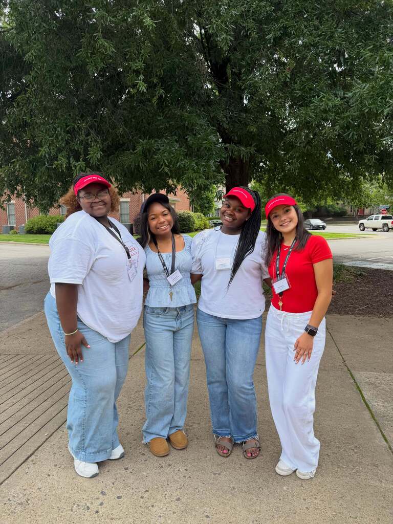 Four individuals stand outdoors on a sidewalk near a large tree. All wear red caps and lanyards. Three wear white tops and jeans; one wears a red shirt and white pants.