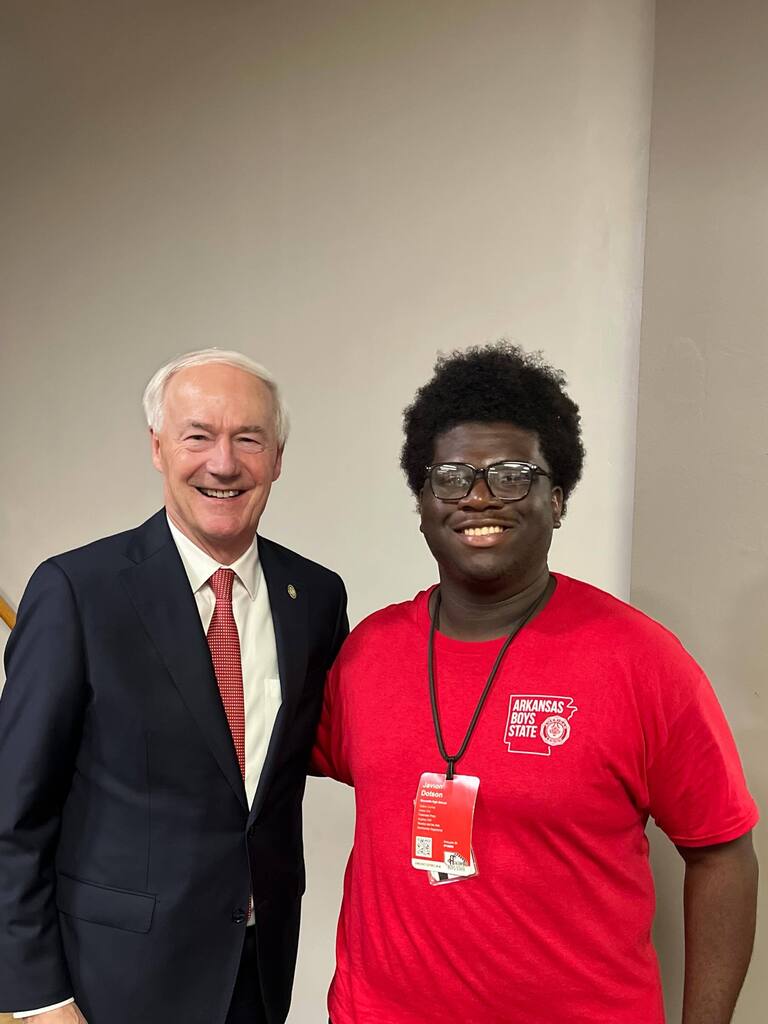 Two individuals stand indoors against a beige wall. One wears a dark suit with a red tie; the other wears a red “Arkansas Boys State” T‑shirt and a red lanyard with a name badge labeled “Dawson.”