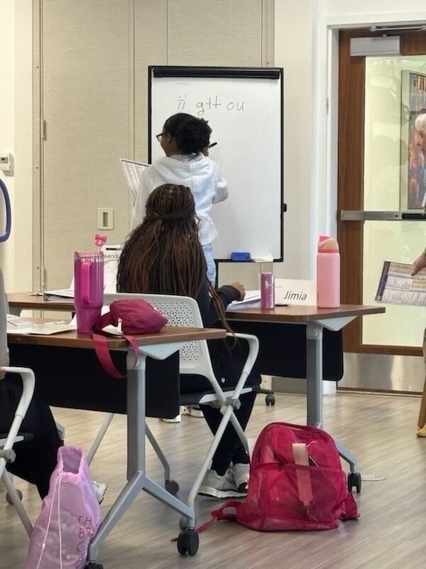 A student stands at a whiteboard writing the phrase “i gtt ou” while classmates sit at desks with name tags, backpacks, and water bottles. The room has light‑colored walls and flooring.