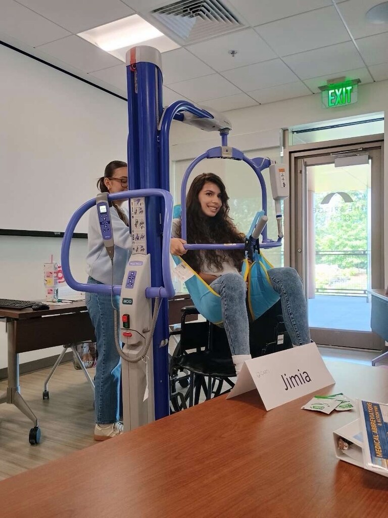 A student operates a mechanical patient lift while another student sits in the attached sling. The classroom includes tables, chairs, and a name card labeled “Jimia” on the table in the foreground.