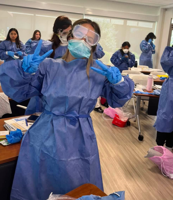 A student in full protective gear—blue gown, gloves, mask, and goggles—stands in a classroom making a peace sign with both hands. Other students in similar gear work at tables covered with papers and supplies.