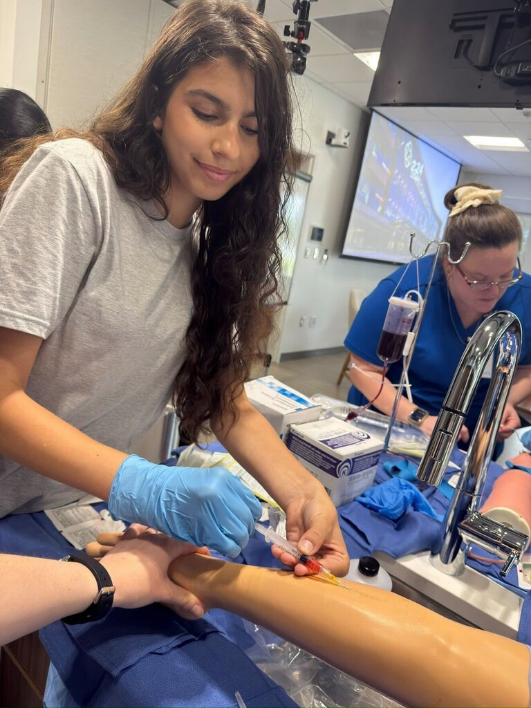 A student wearing blue gloves inserts a syringe into a training arm while another student in scrubs works with supplies nearby. The room includes medical equipment, boxes of materials, and a large wall‑mounted monitor.