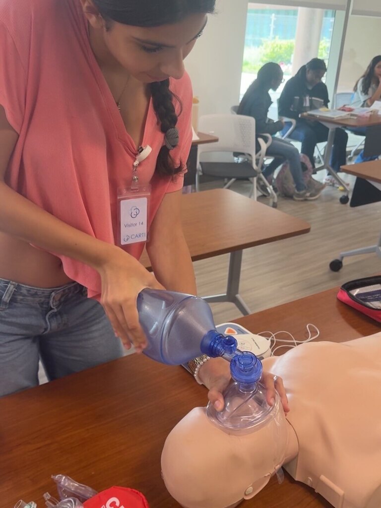 A student wearing a visitor badge labeled “Visitor 14” practices using a bag‑valve mask on a CPR manikin placed on a wooden table. Other participants sit at desks in the background.
